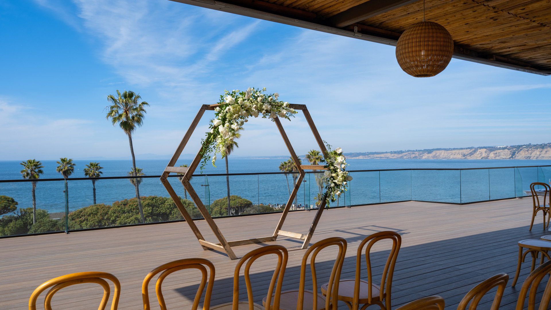 Modern geometric wooden wedding arch with white floral arrangements overlooking Pacific Ocean and La Jolla coastline, featuring bentwood ceremony chairs at Cove Rooftop venue