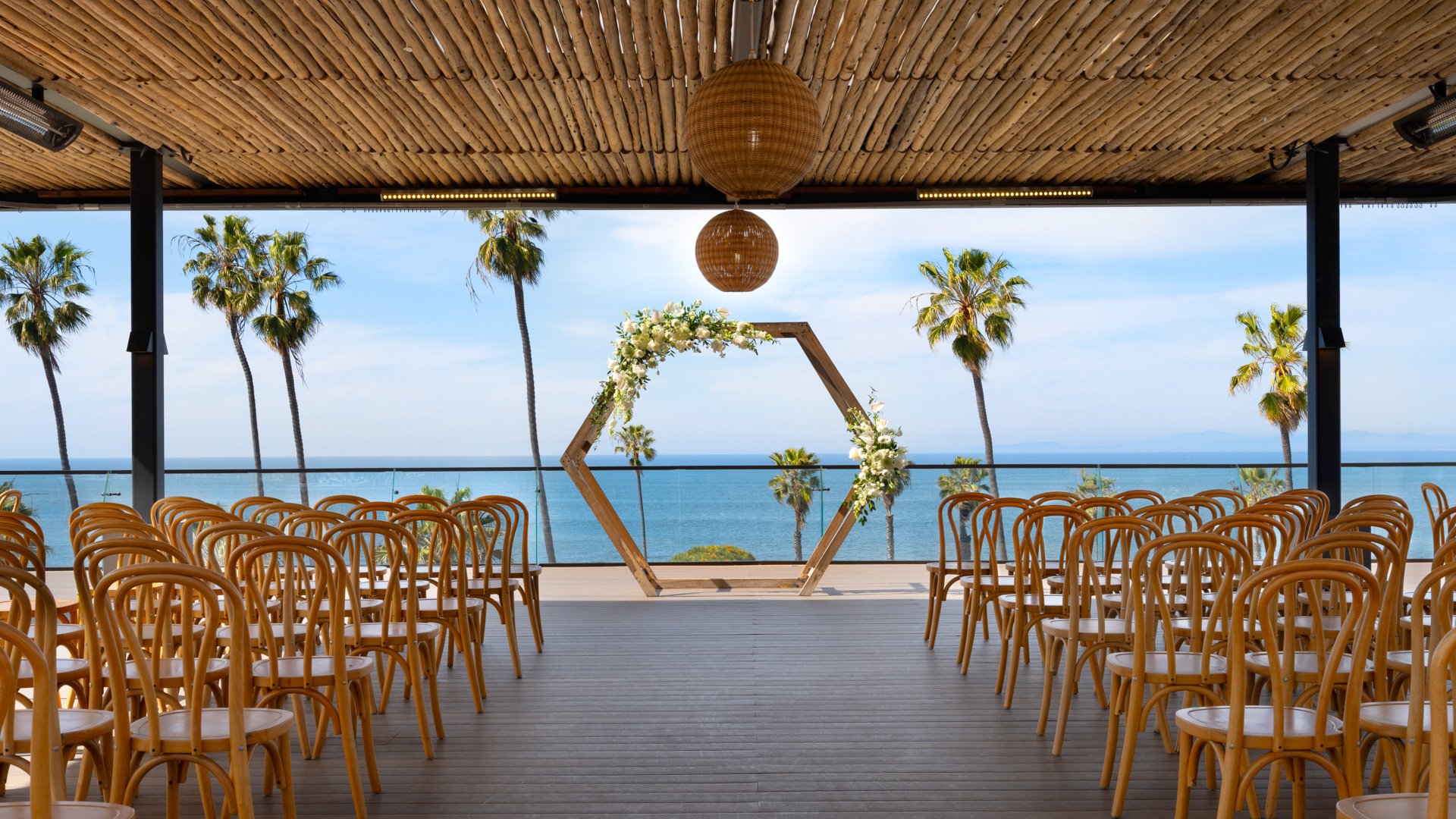 Beachfront wedding ceremony space at La Jolla Cove Rooftop featuring geometric floral arch, bentwood chairs, and panoramic Pacific Ocean views with palm trees