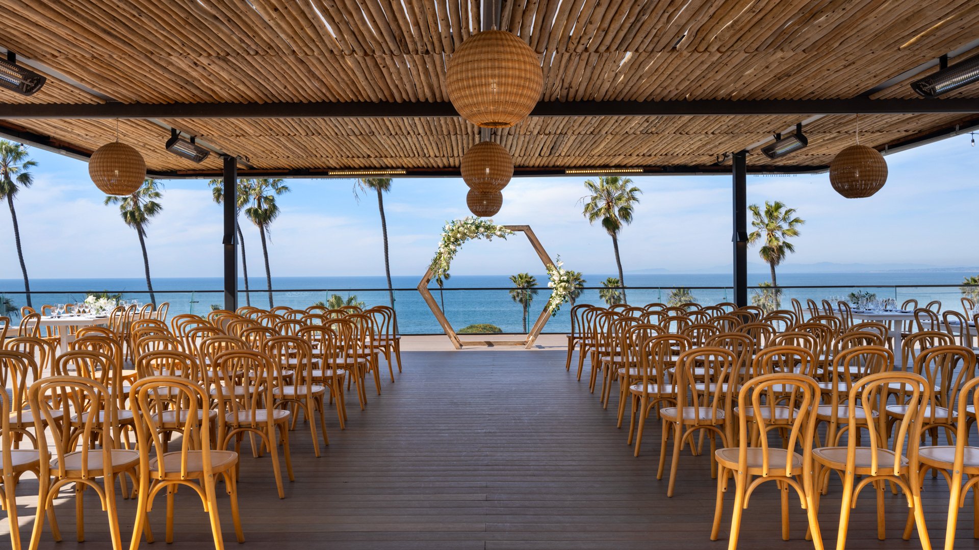 Spacious wedding ceremony with bentwood chairs, geometric floral arch, and woven pendant lights under natural wood pergola at La Jolla Cove Rooftop by Wedgewood Weddings