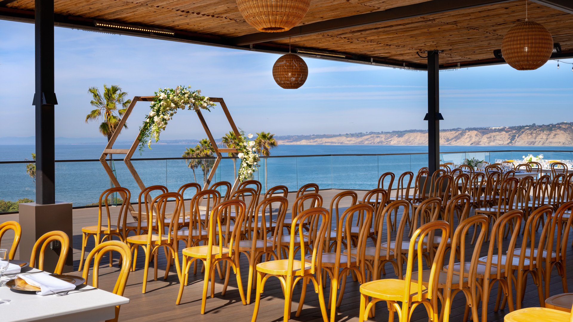 Beachfront wedding ceremony setup with geometric wooden arch adorned with white florals, bentwood chairs, and Pacific Ocean backdrop at La Jolla Cove Rooftop