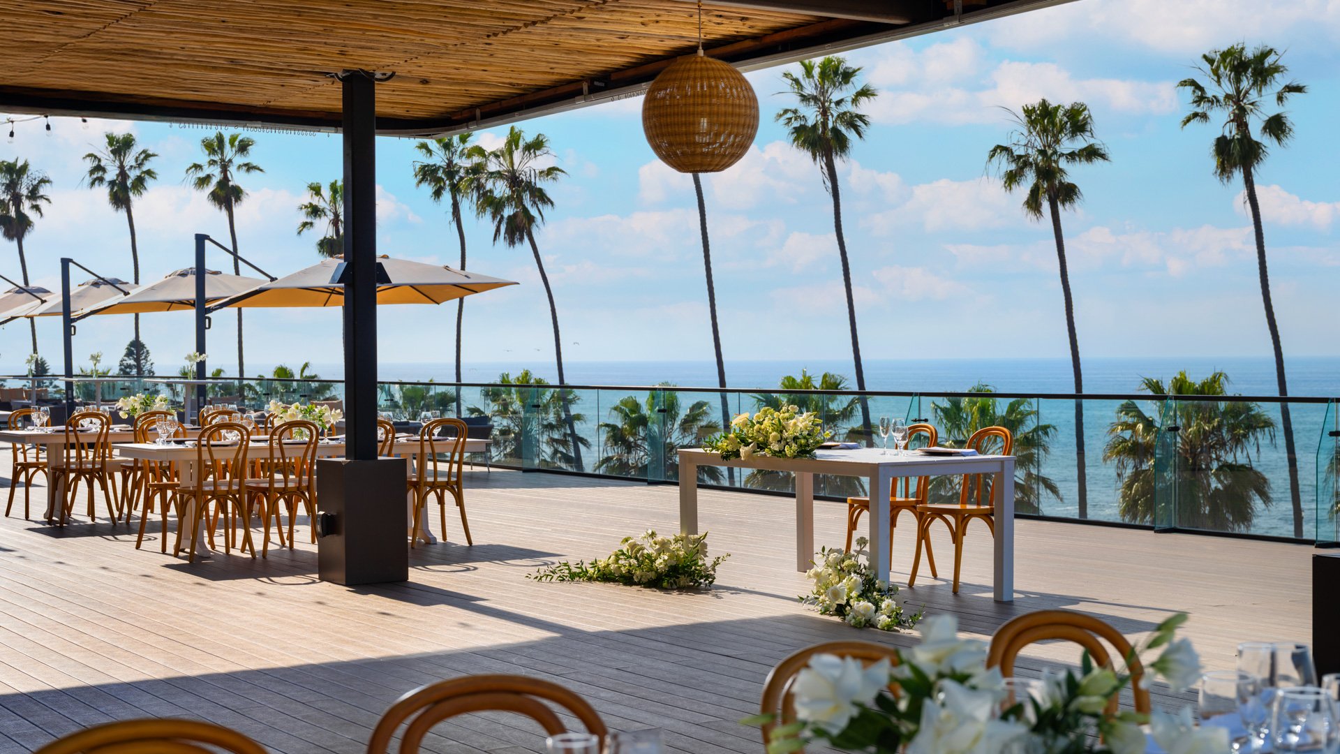 Large outdoor wedding reception terrace with bentwood chairs, natural wood ceiling, and tropical palm tree landscape at La Jolla Cove Rooftop