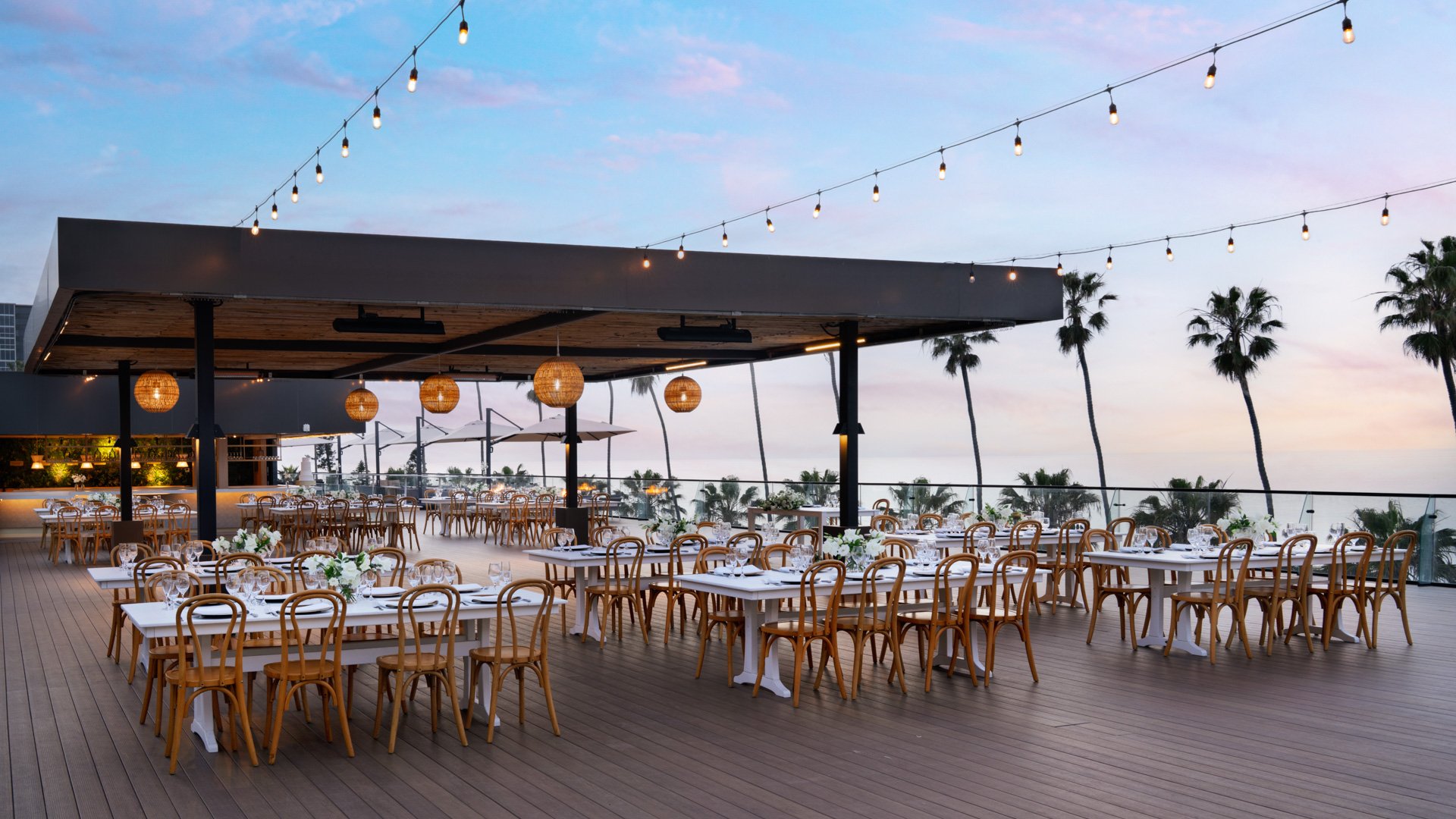 Romantic evening wedding reception under string lights with bentwood chairs, white tables, and natural wood pergola at La Jolla Cove Rooftop by Wedgewood Weddings