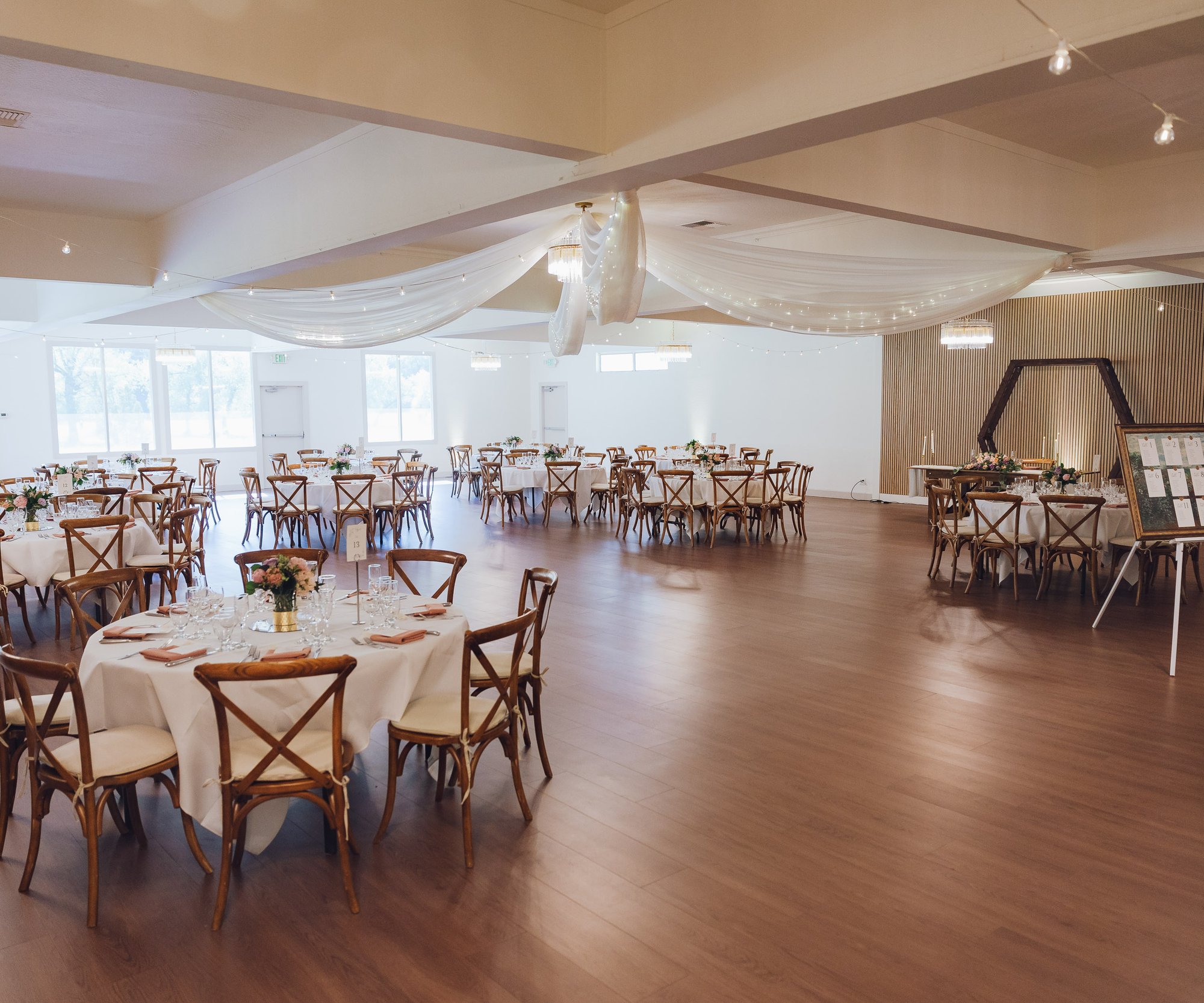 Large reception hall with multiple round tables, cross-back chairs, and dramatic crystal chandelier under white ceiling draping
