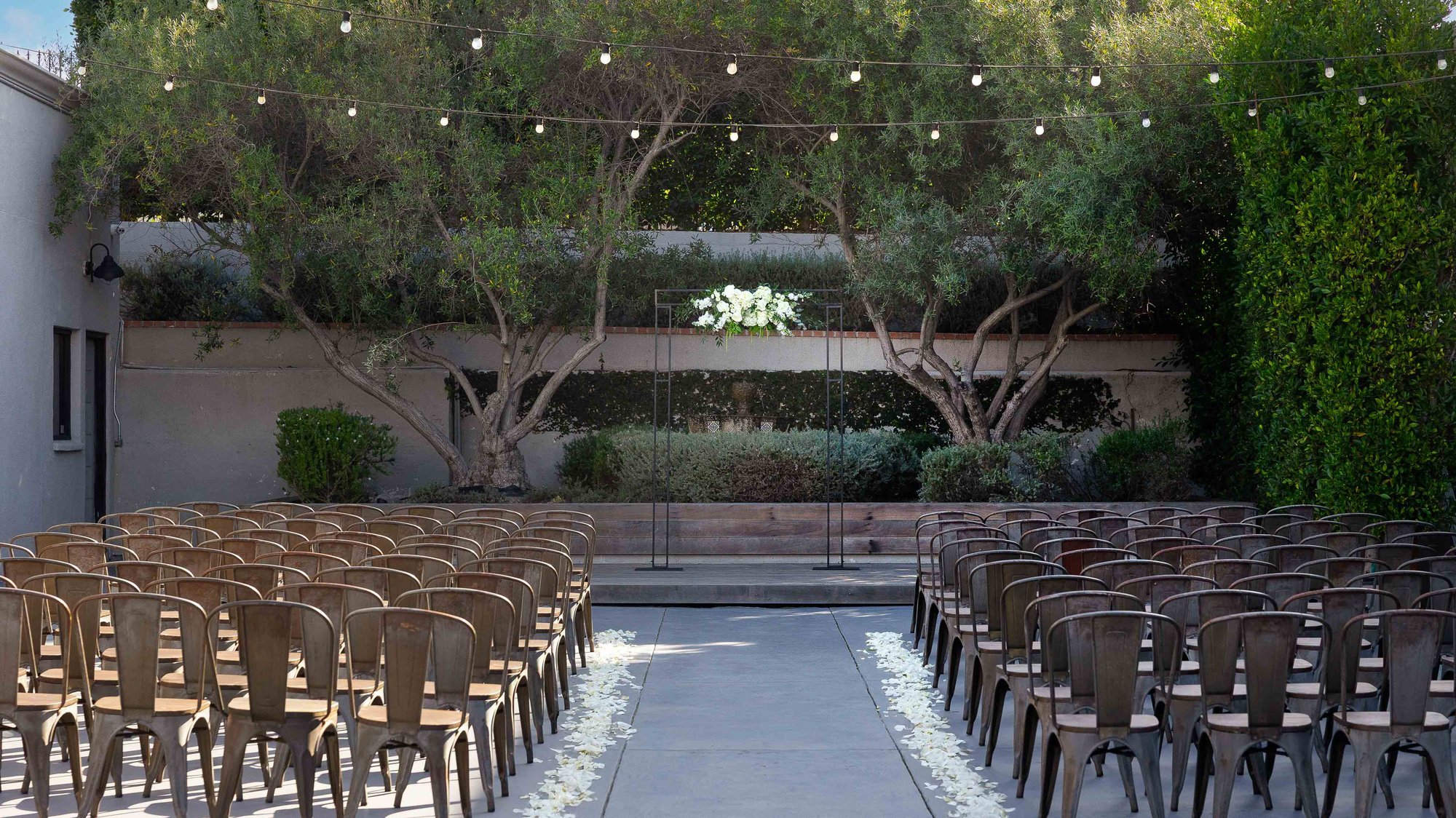 An outdoor ceremony space with rows of metal chairs, string lights overhead, and a floral altar arrangement