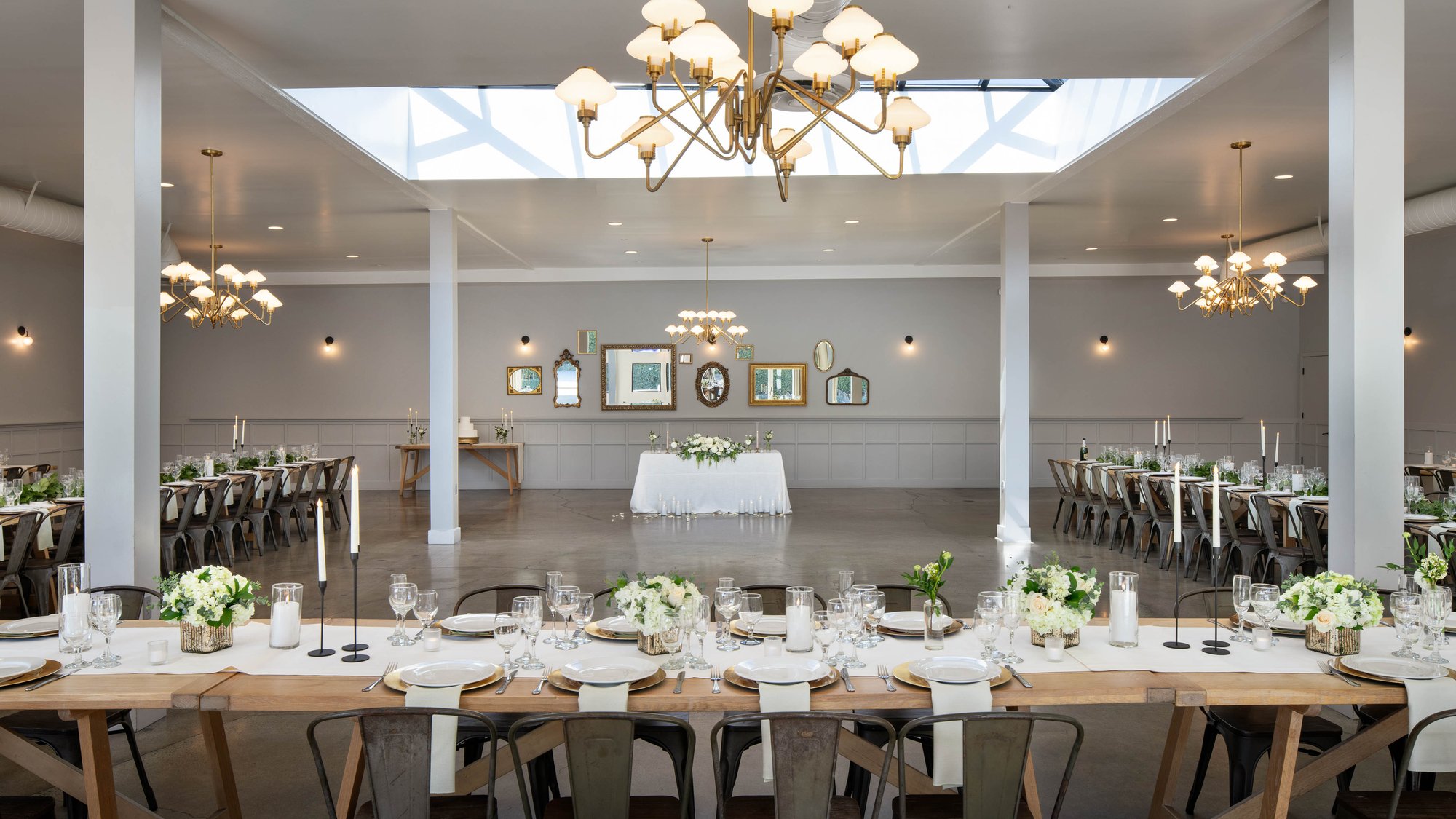 An elegant indoor reception hall with gold chandeliers, white tablecloths, and farmhouse-style tables